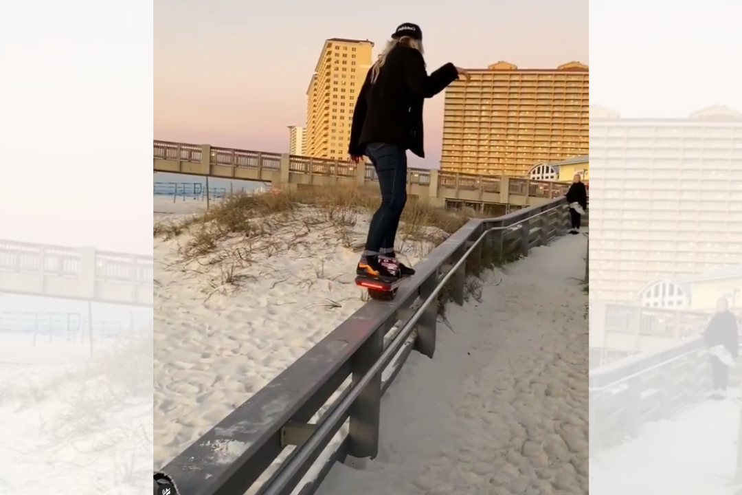 Screenshot of Kristy Miller riding her onewheel up a 5 inch wide fence, 6 feet in the air on the beach with hues of orange, brown, blue, and black in the landscape.